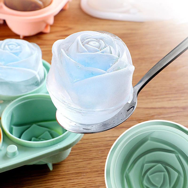 A light blue colored rose shaped ice cube is help in a spoon, behind this is a green colored rose shaped ice cube mold maker.