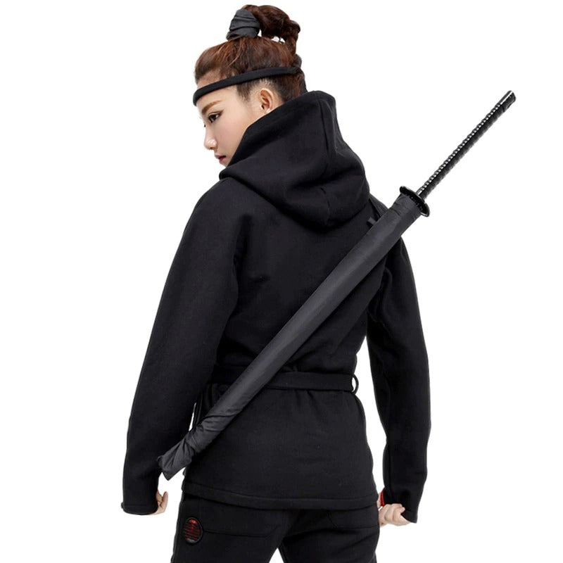 A female ninja dressed in black with a headband carrying a samurai sword umbrella slung over her shoulder.