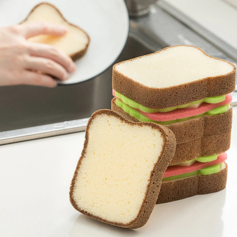 A sponge which lots like a piece of toast. There is also a sponge which looks like a fully loaded sandwich complete with lettuce. The sponges are resting on a kitchen sink.