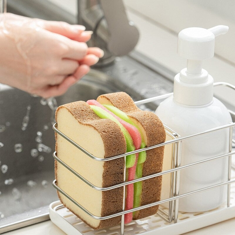 A sponge which looks like a complete sandwich with fillings. The sponge is sitting in a sponge holder on a kitchen sink.