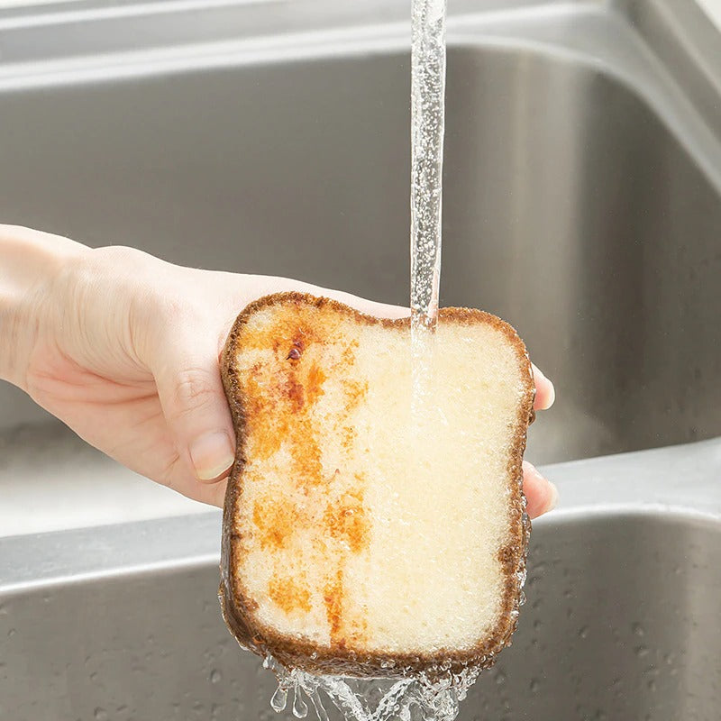 A sponge which looks like a slice of bread. It is being held under a running tap and half of the slice looks dirty while the side under the running water is clean.