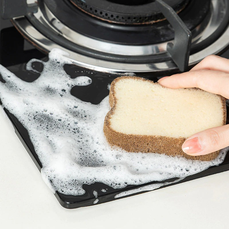 A hand is using a bread sponge to clean around a stovetop. The sponge looks like a slice of bread.