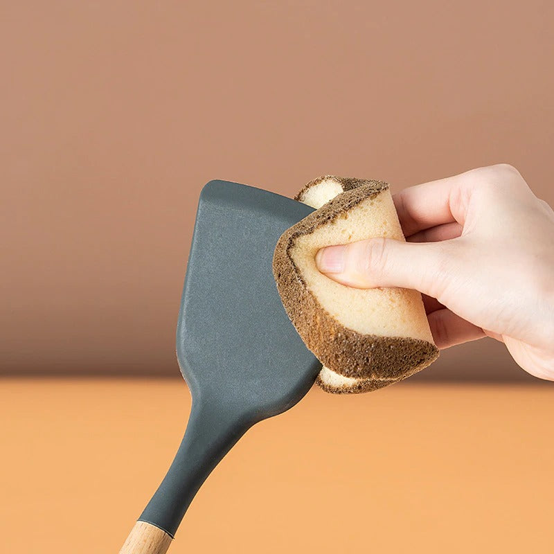 A bread sponge is cleaning a grey spatula with a wooden handle. A bread sponge is a sponge shaped like a piece of bread.