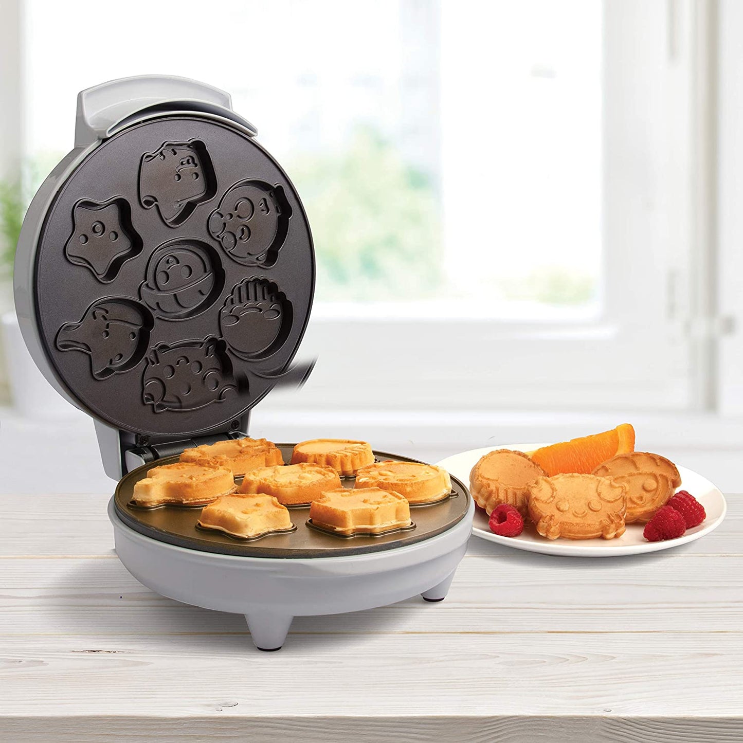 An electric waffle maker which makes sea creature shaped waffles for kids. There are cooked examples in the maker as well as on a white plate next to the waffle maker. They look delicious.