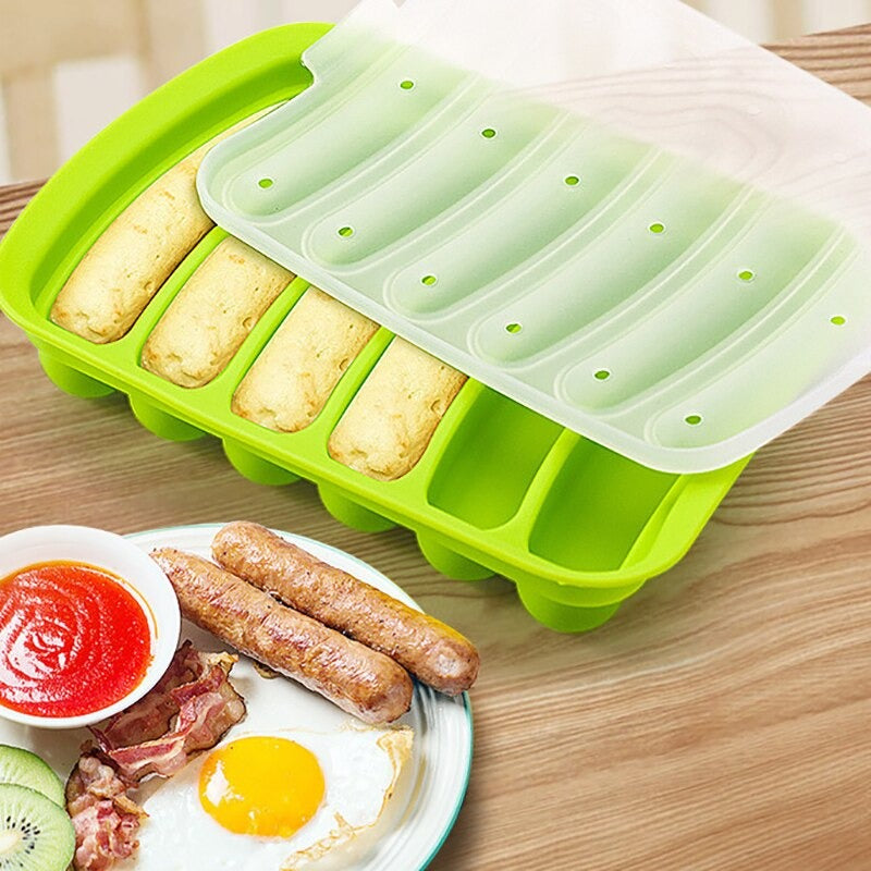 A green silicone sausage mold with 4 sausages inside the mold and a plate of food next to the mold which includes 2 cooked sausages, eggs, bacon and tomato sauce.