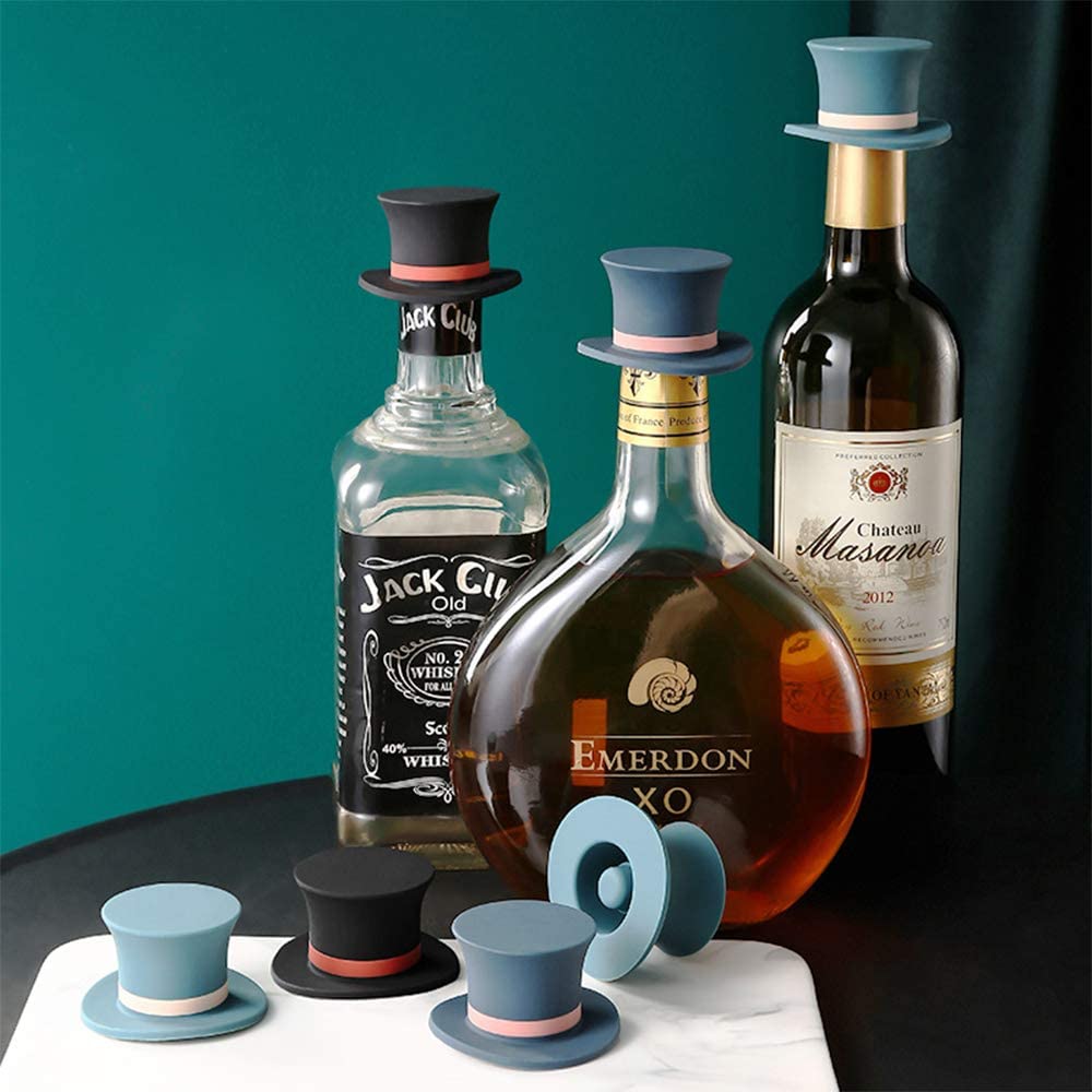 Four different colored silicone wine bottle stoppers shaped as top hats resting in front of 3 different types of alcohol bottles.