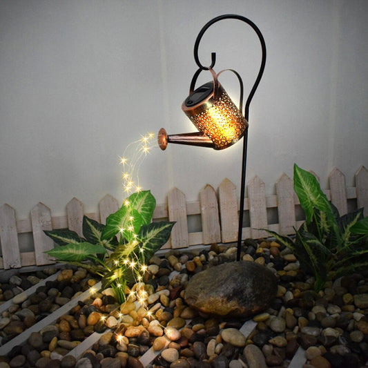 A garden décor item which is a small solar powered watering can with fairy lights coming out of the spout in a garden at night.