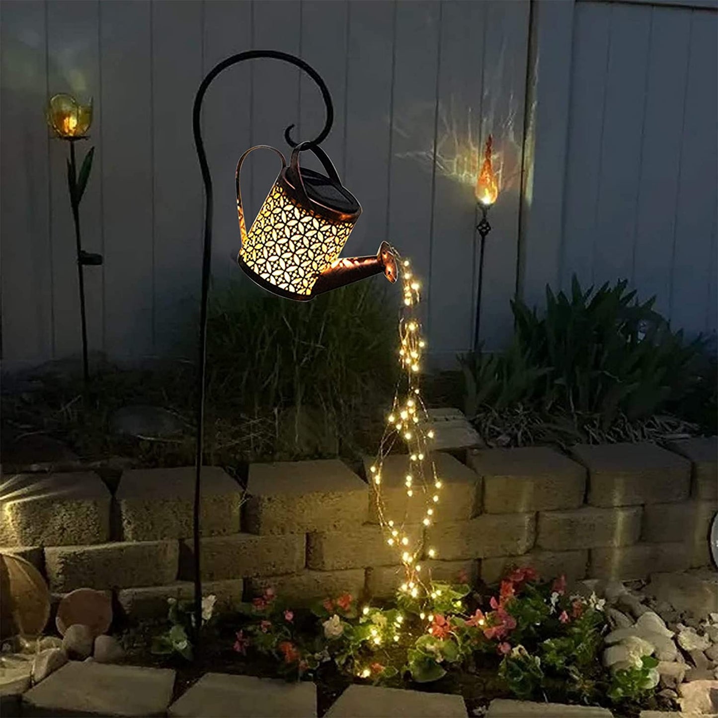 A garden décor item which is a small solar powered watering can with fairy lights coming out of the spout in a garden at night.