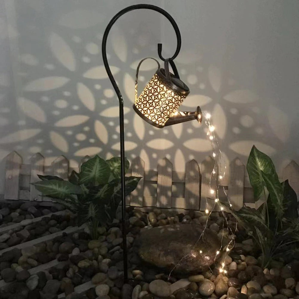 A garden décor item which is a small solar powered watering can with fairy lights coming out of the spout in a garden at night.
