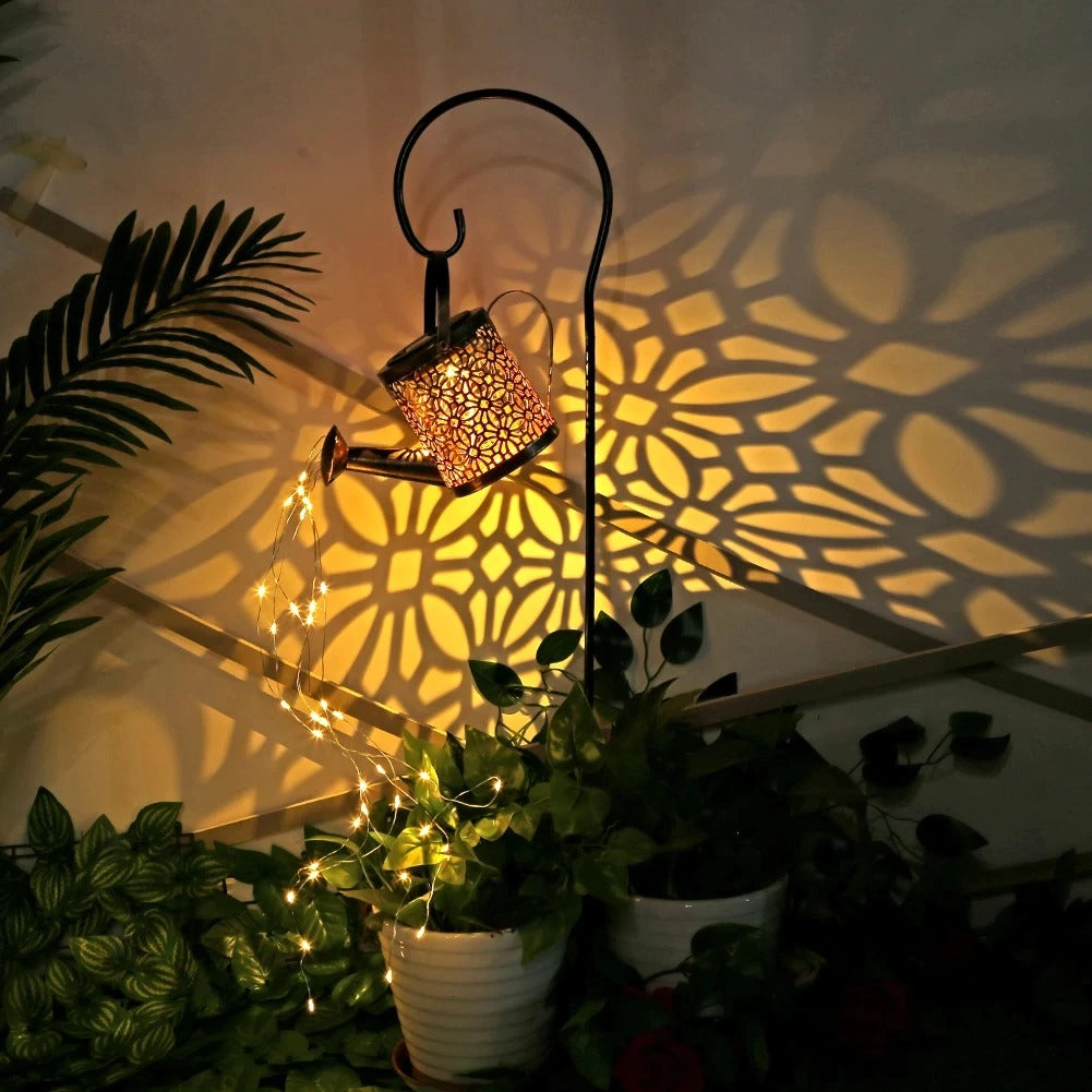 A garden décor item which is a small solar powered watering can with fairy lights coming out of the spout in a garden at night with some pot plants in the background.