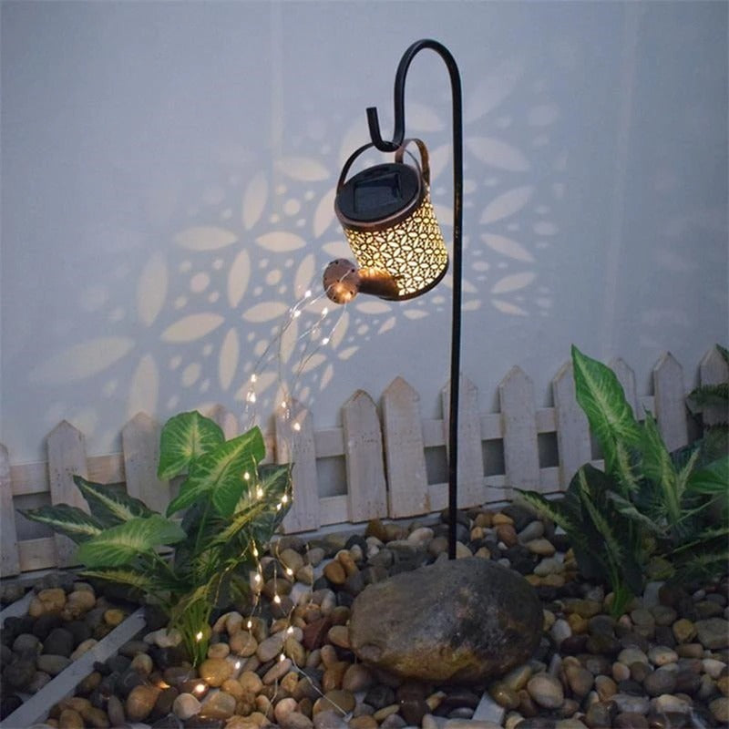 A garden décor item which is a small solar powered watering can with fairy lights coming out of the spout in a pebbled garden at night.