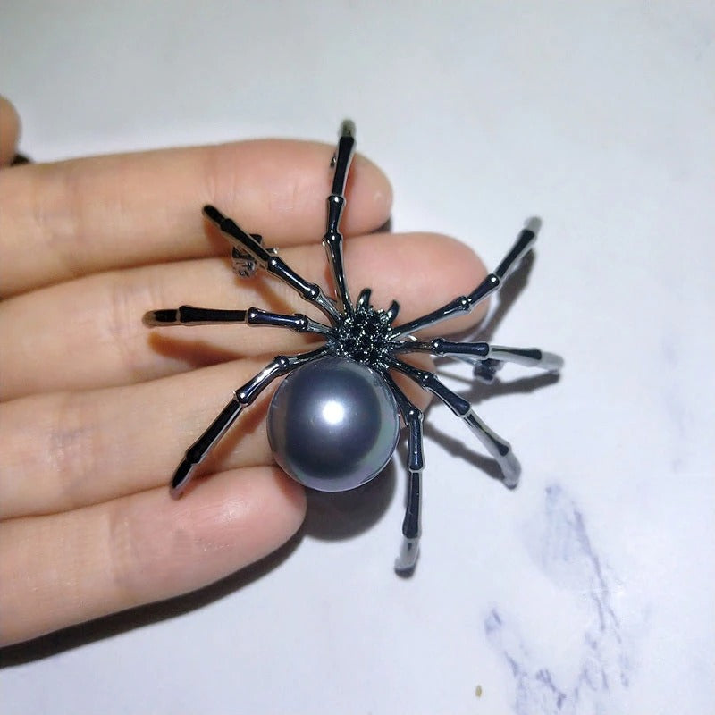 A black spider brooch with small faux black gems and a simulated black pearl. The brooch is on the fingertips of a palm showing its size and details closeup. The spider brooch appears to be falling off the fingers onto a marble table.