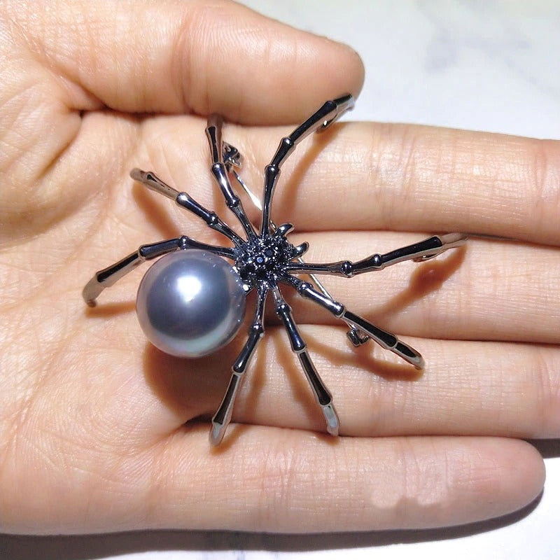 A black spider brooch with small faux black gems and a simulated black pearl. The brooch is on the fingertips of a palm showing its size and details closeup.