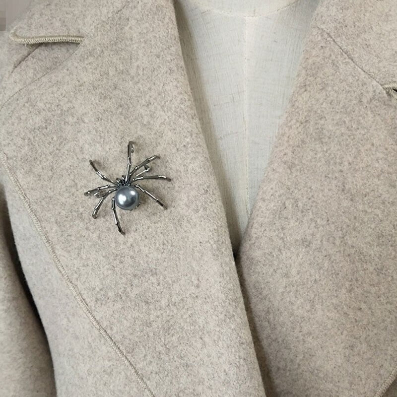 A black spider brooch with small faux black gems and a simulated black pearl. The brooch is being worn by a person and is shown on the lapel of a jacket.