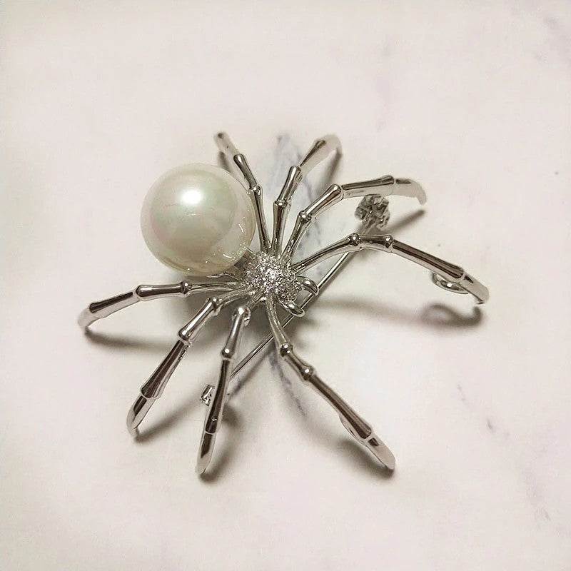 A silver colored spider brooch with a white simulated pearl sitting on a white background. 