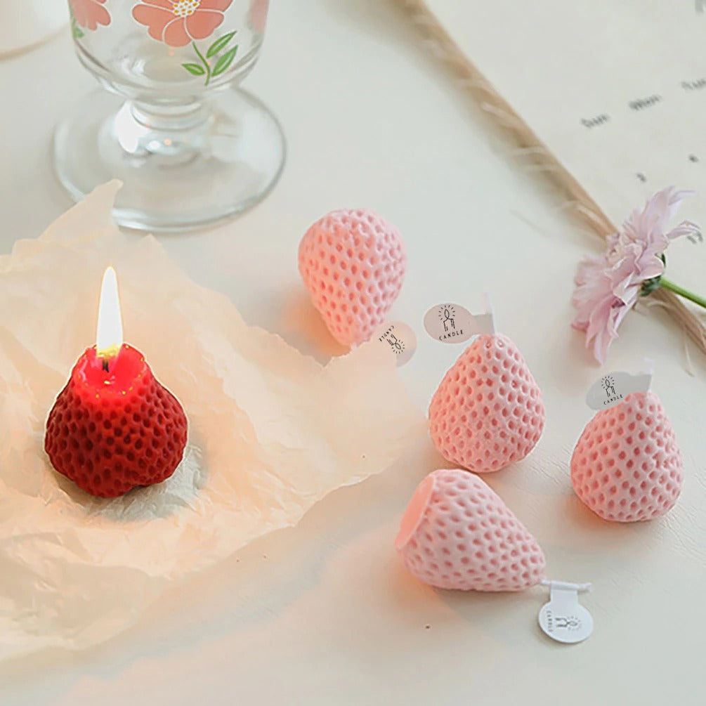 One red strawberry shaped candle and four pink strawberry shaped candles. The red strawberry candle is alight and two of the pink ones are laying on their side.