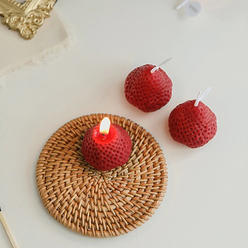 Three red strawberry shaped candles, one is alight on a bamboo coaster and the other two red candles are off to the side.