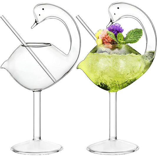 Two swan shaped cocktail glasses each with a glass straw. One of the glasses as a green drink in it with a mint and floral garnish.