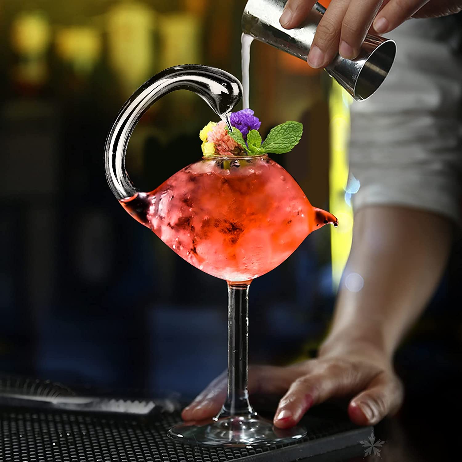 A hand is pouring liquid from a shot glass into a swan shaped cocktail glass. The glass is filled with a red drink with a mint and floral garnish.