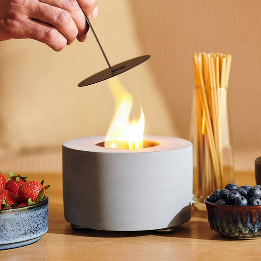 A white indoor/outdoor table top fireplace with a flame coming out of the centre. A hand is about to put a lid on to extinguish the flame.