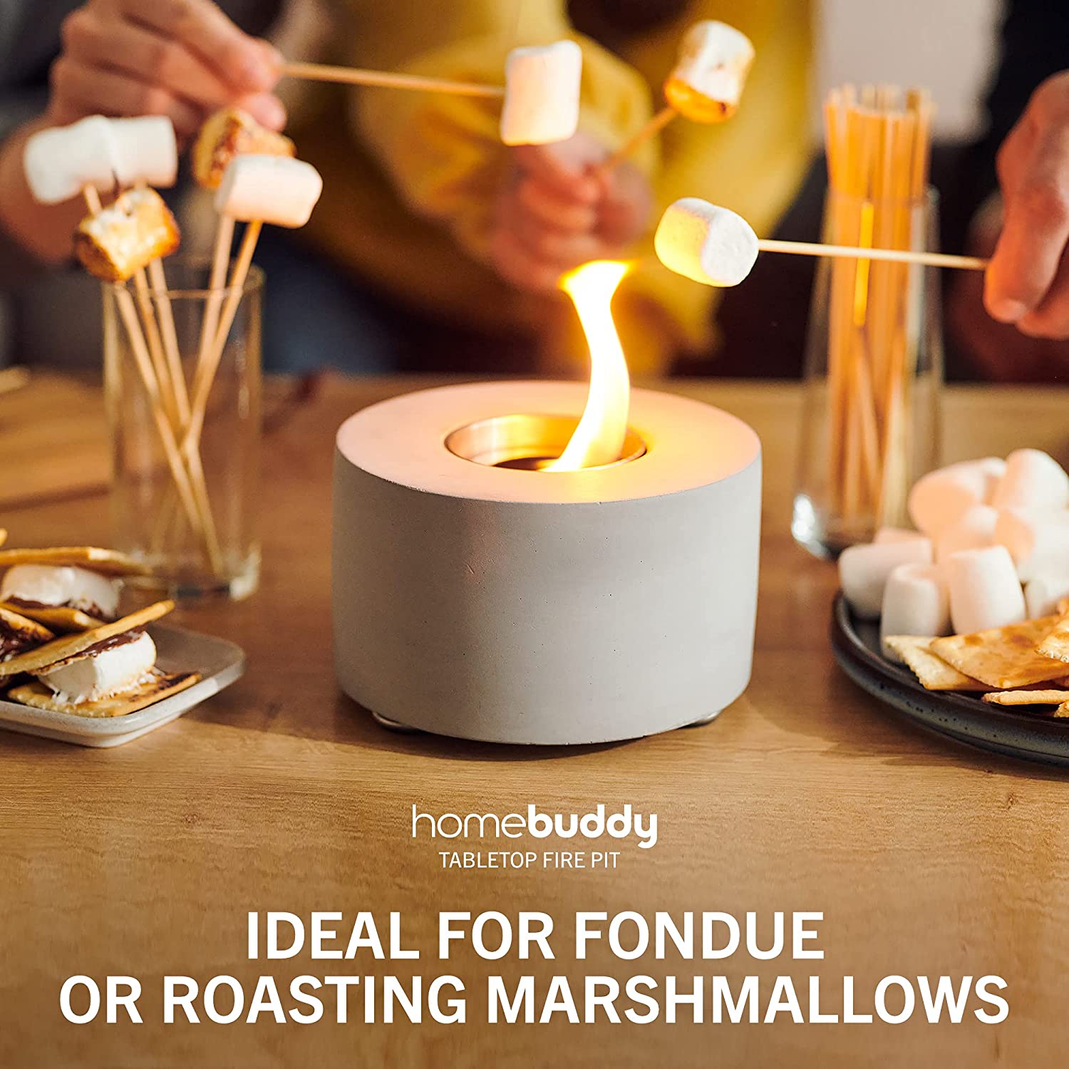 A group of people are roasting marshmallows over a small indoor table top fire pit. There is a plate of S'mores nearby.