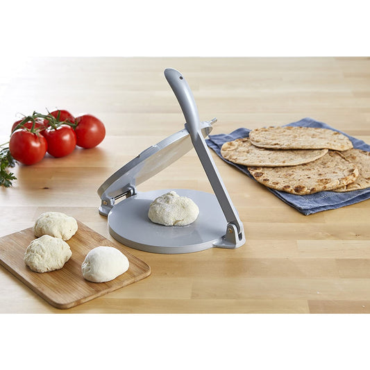 A silver colored aluminium tortilla press with fresh dough on the press plate. Nearby are some freshly cooked tortillas and some tomatoes.