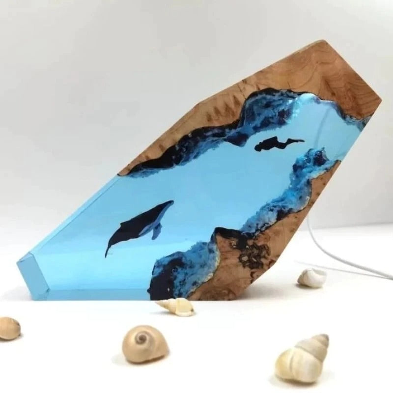 A desk lamp which looks like a slice of underwater life. There is a diver and a humpback whale swimming toward each other in an underwater cave. The light itself is not switched on. There are 5 sea shells used as decoration near the lamp.