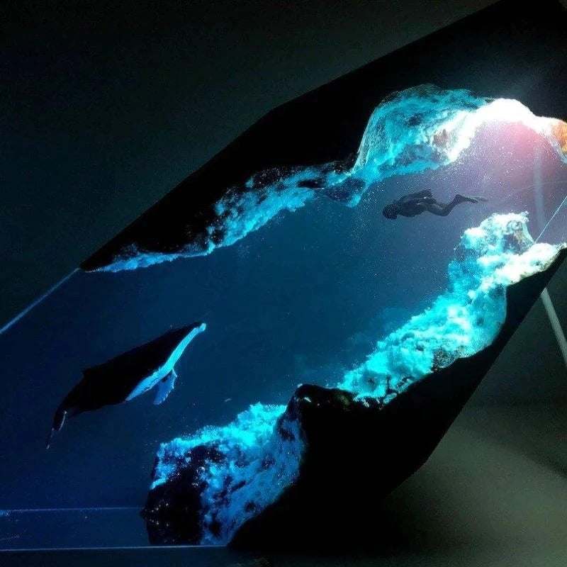 A closeup of a lamp which features an underwater scene. There is a cave with a diver swimming towards a humpback whale. The lamp is switched on and illuminates the scene in a blue light.