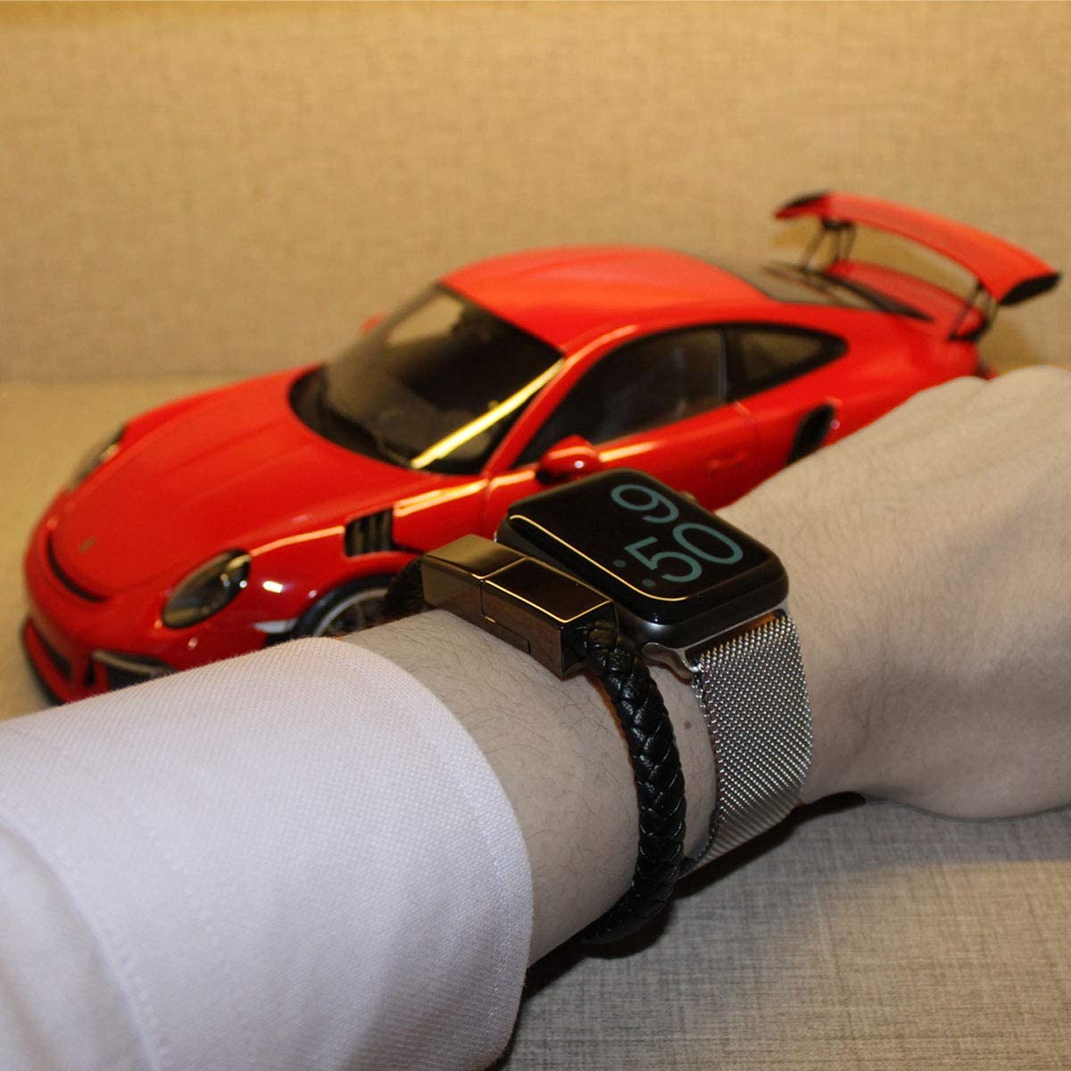 A person is wearing a black leather USB charging bracelet on his wrist along with a black watch. There is a red model car in the background.