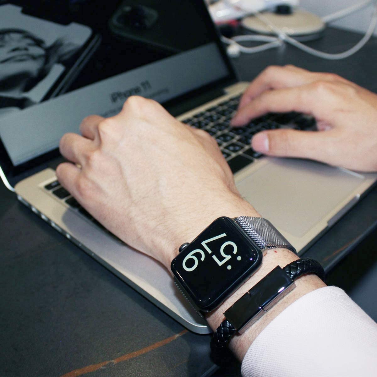 A person has their hands and wrists resting on a laptop keyboard. The person is wearing a USB charging bracelet along with a black watch.