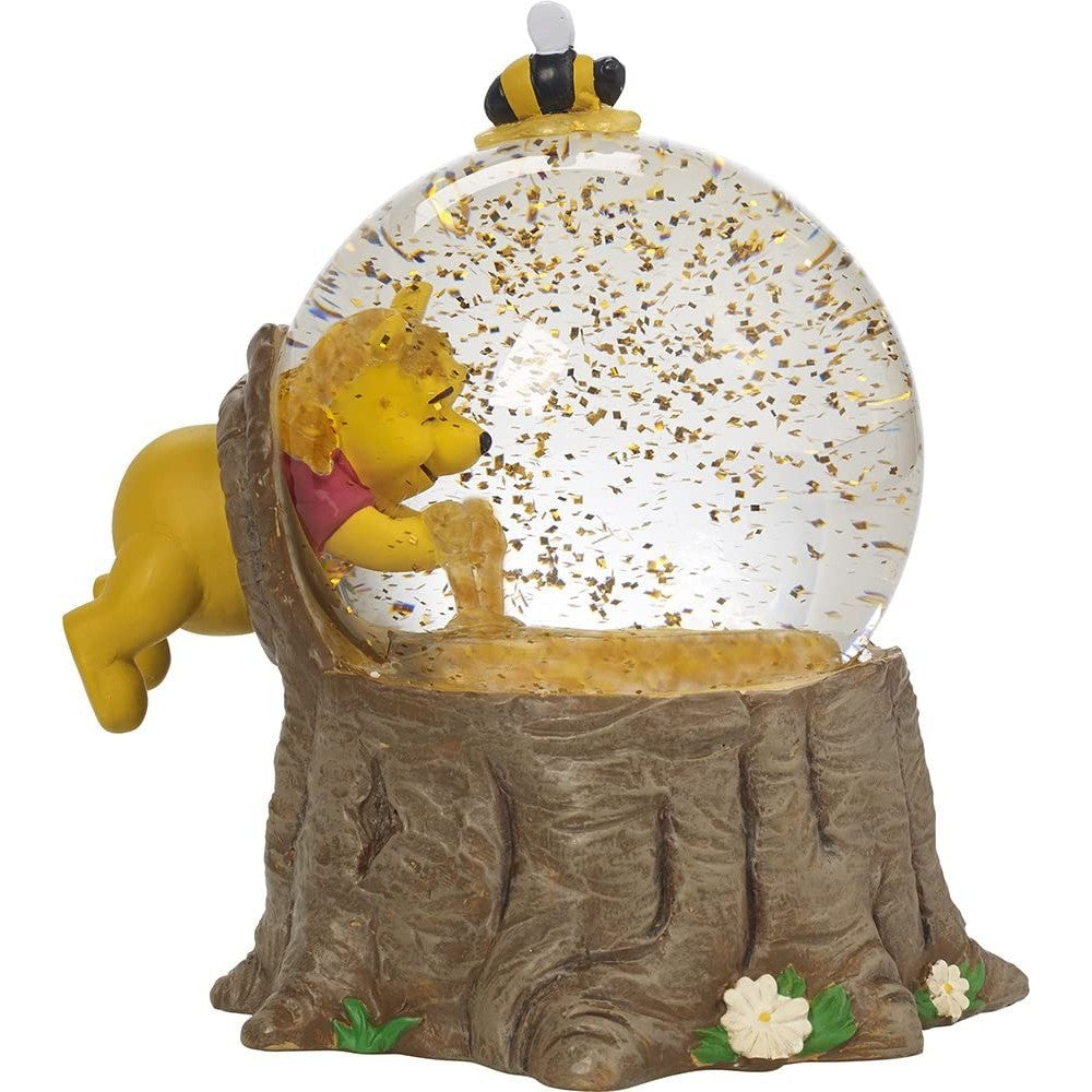 A Winnie the Pooh snow globe depicting Winnie putting his hands in a hive full of honey. There is glitter swirling around which look like bees