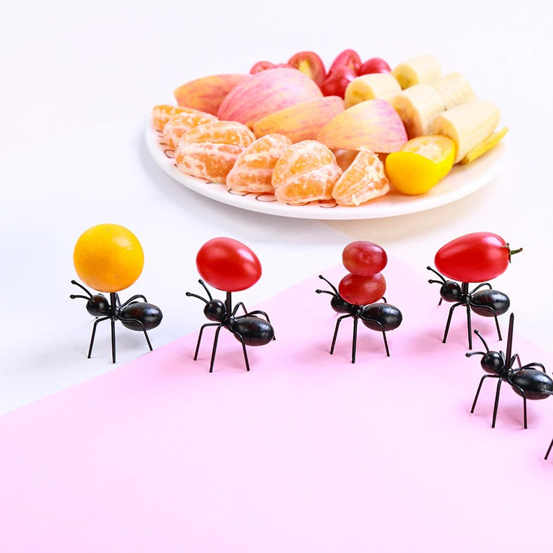 5 plastic worker ant food picks marching on a pink and white background. In the foreground some of the ants are carrying grapes and cherry tomatoes while a plate of fruit is in the background.