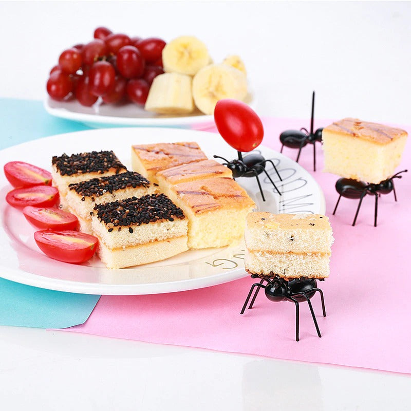 4 plastic worker food ant picks. One is carrying a cherry tomato while 2 others are carrying slices of cake and one is carrying nothing. There is one plate with cakes and another plate in the background with sliced banana and red grapes.
