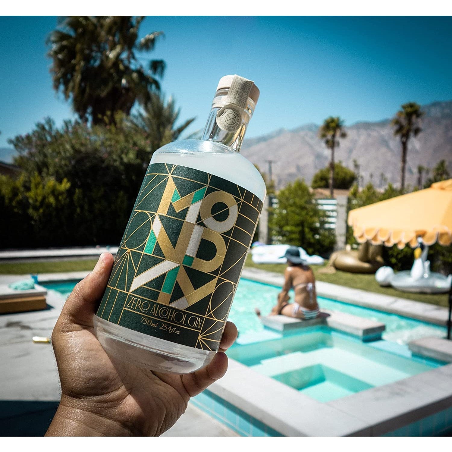 A hand is holding a bottle of Monday non-alcoholic gin in front of a swimming pool on a sunny day.